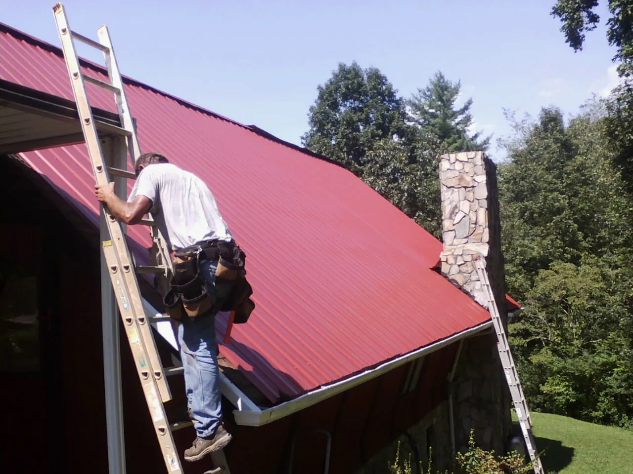 Roofer on a ladder beside a red metal roof and stone chimney — Soffit & Fascia Repair by Roofing Brothers in Sabinsville